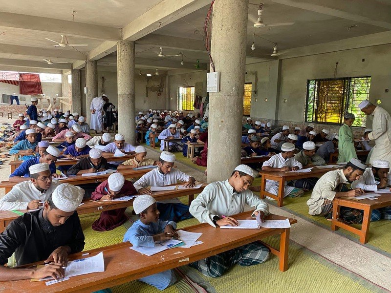 Students writing examination papers at benches in a large hall